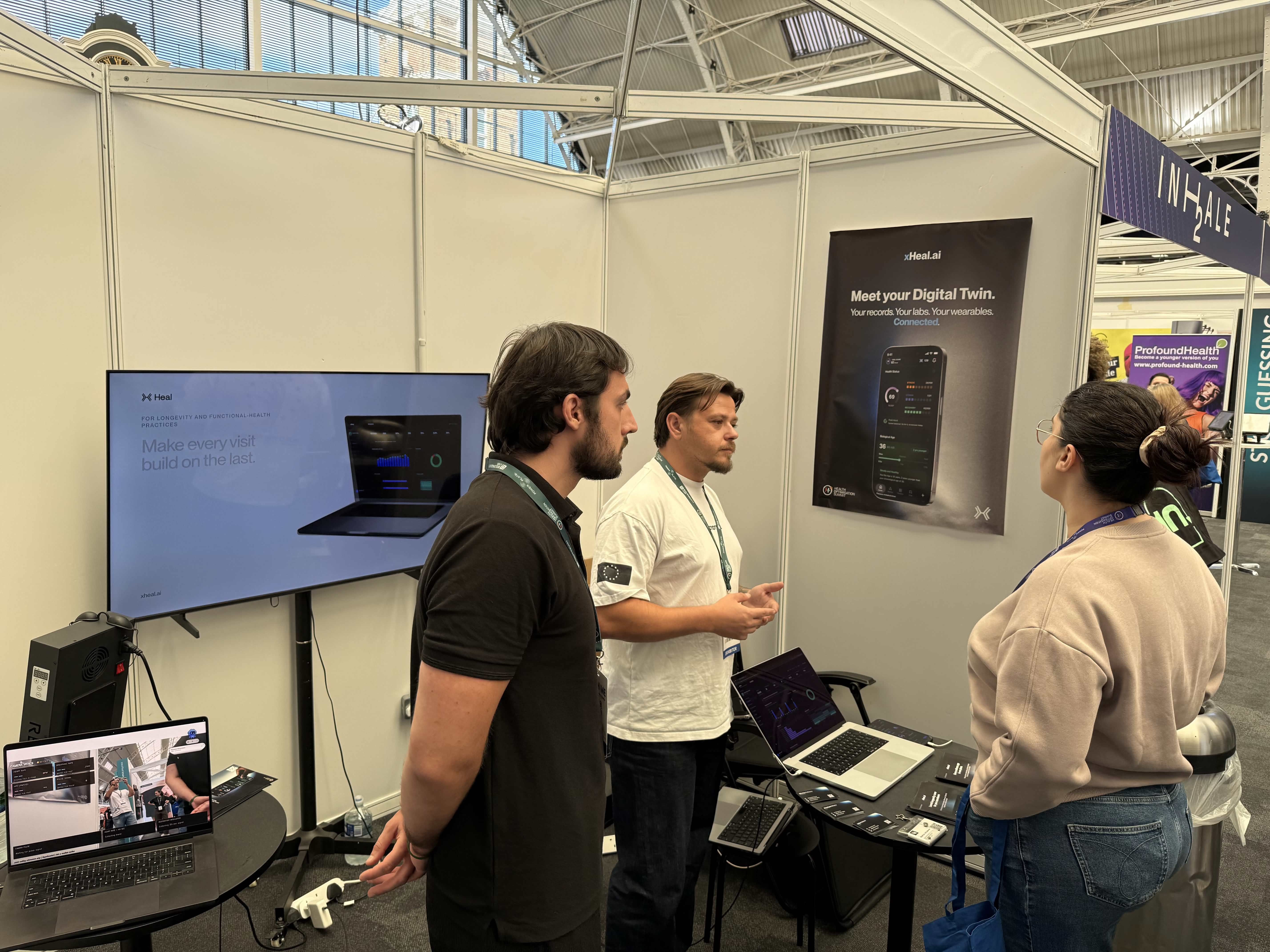 Two xHeal team members talking with an attendee at the booth, beside a screen reading 'Make every visit build on the last' for longevity and functional-health practices