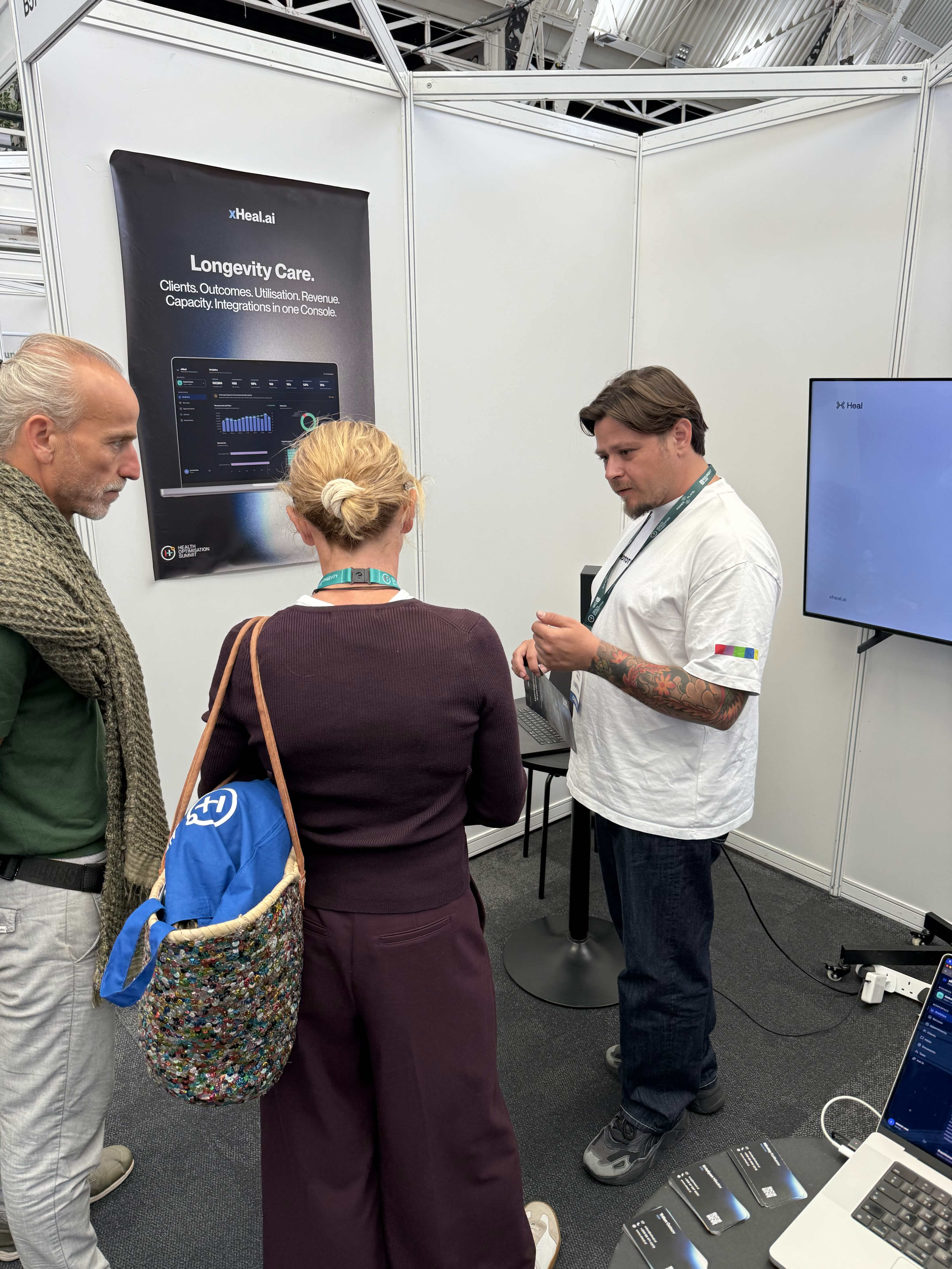 An xHeal team member demonstrating Provider Workspace to two attendees, beside the Longevity Care console poster