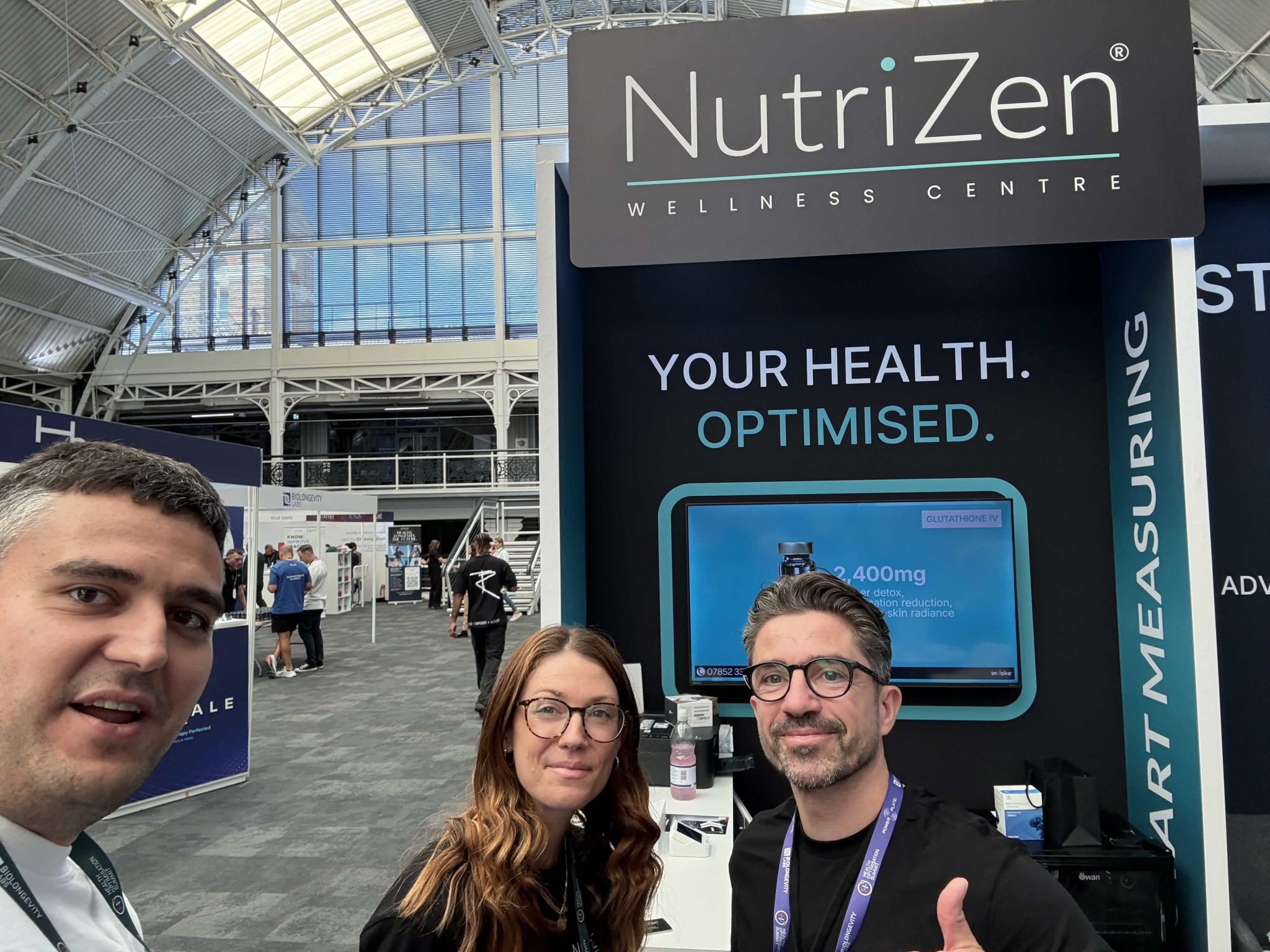 An xHeal team member with two visitors at the NutriZen Wellness Centre stand, showing an IV therapy screen
